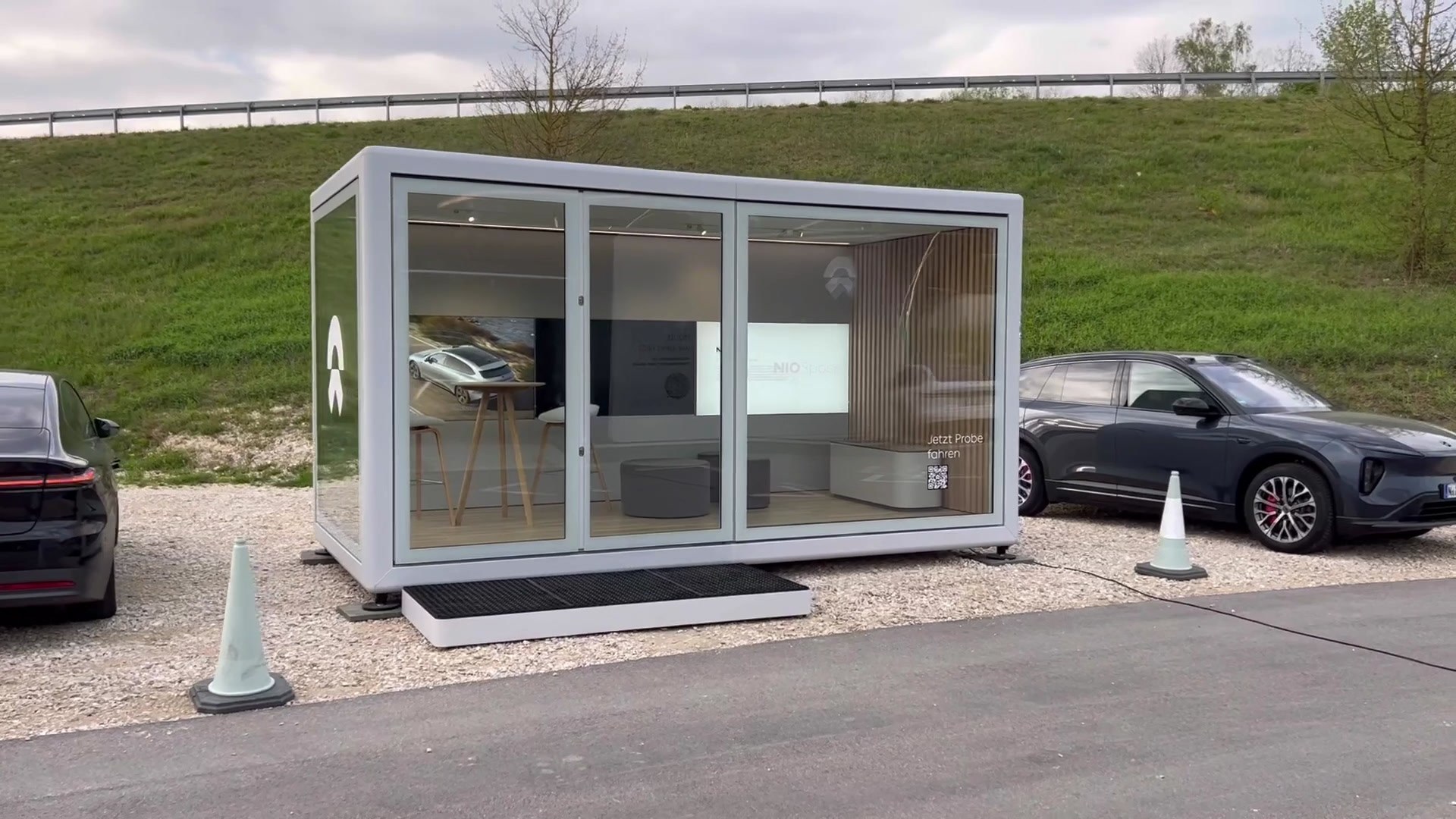 Weißes KOYA Roadshow-Modul mit Glasfront und Eingangsstufe vor einer Böschung, innen eine Ausstellung, daneben zwei Fahrzeuge