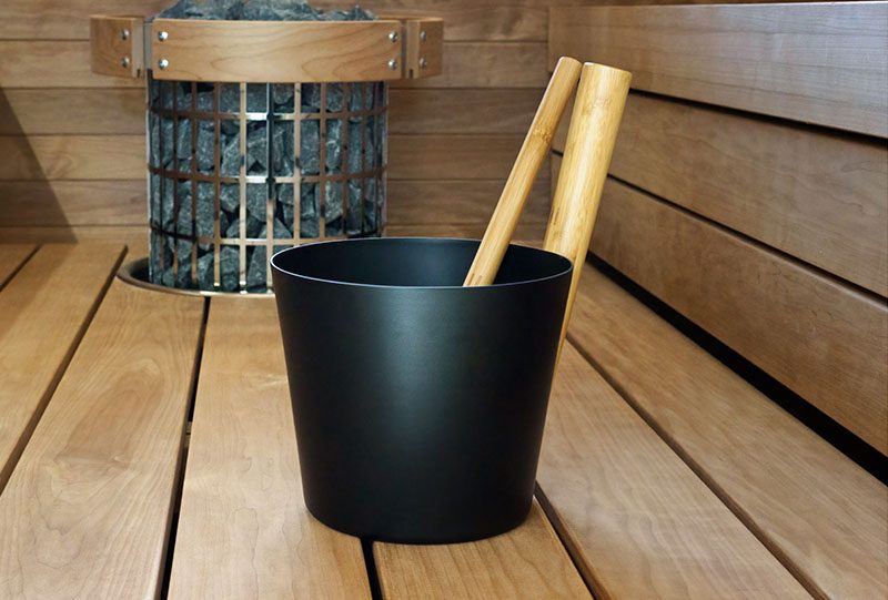 Schwarzer Aufgusseimer mit Kellenstielen aus Holz auf der Holzbank einer Sauna, dahinter ein Ofen mit Steinen