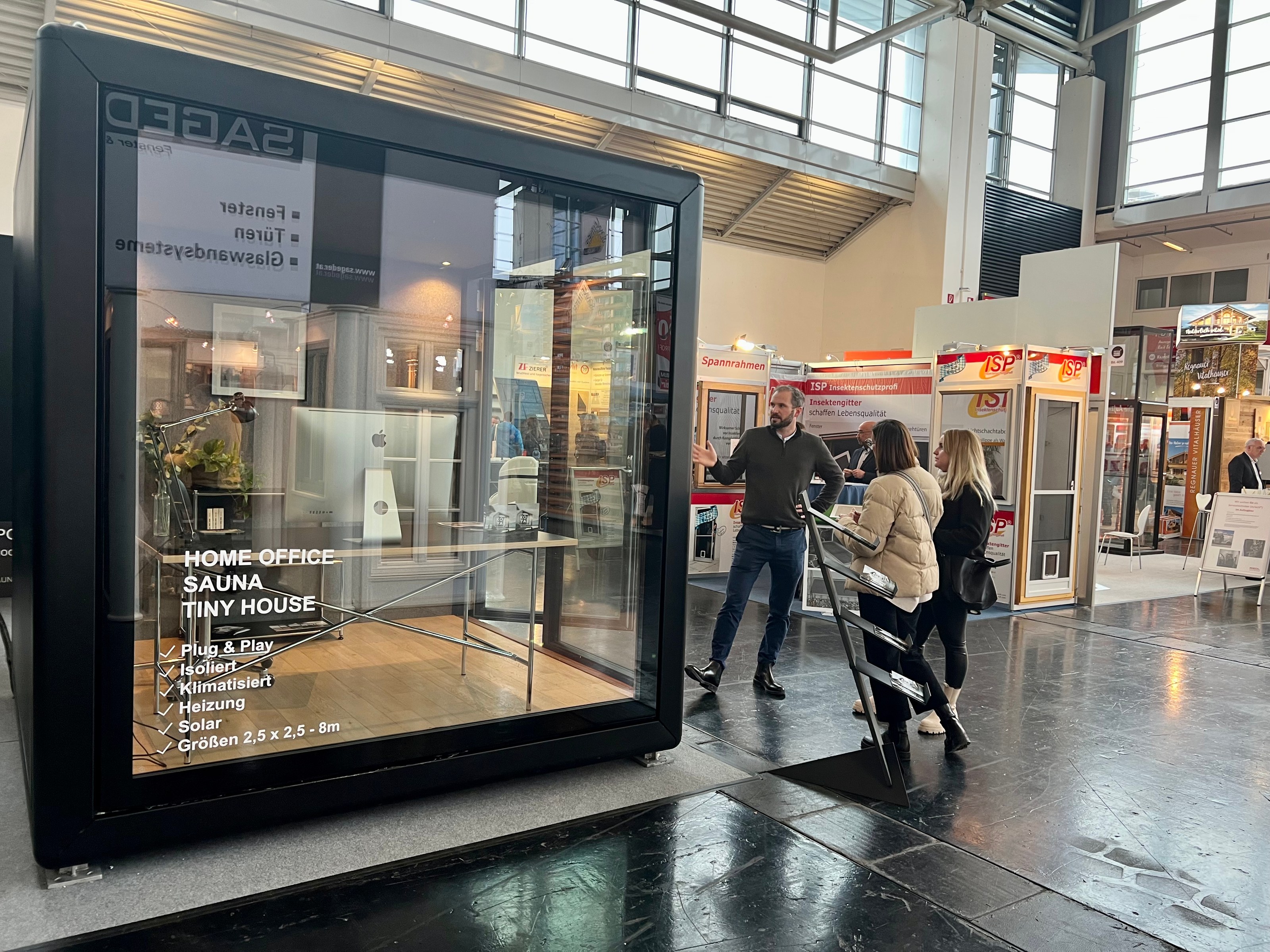 Schwarzer Glaswürfel auf einer Messe mit Holzboden und Schreibtisch; Schriftzug auf der Scheibe nennt Home Office und Sauna.
