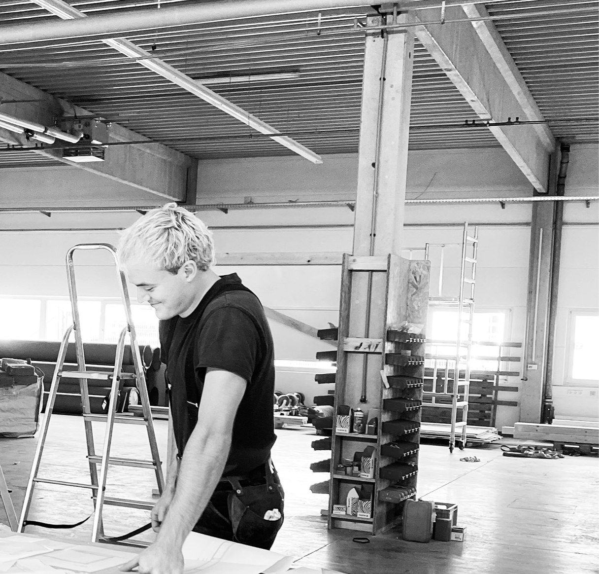 Schwarzweißfoto: Mann in dunklem Shirt beim Arbeiten an Unterlagen auf einem Tisch in einer Werkhalle mit Leitern und Werkzeugregal.