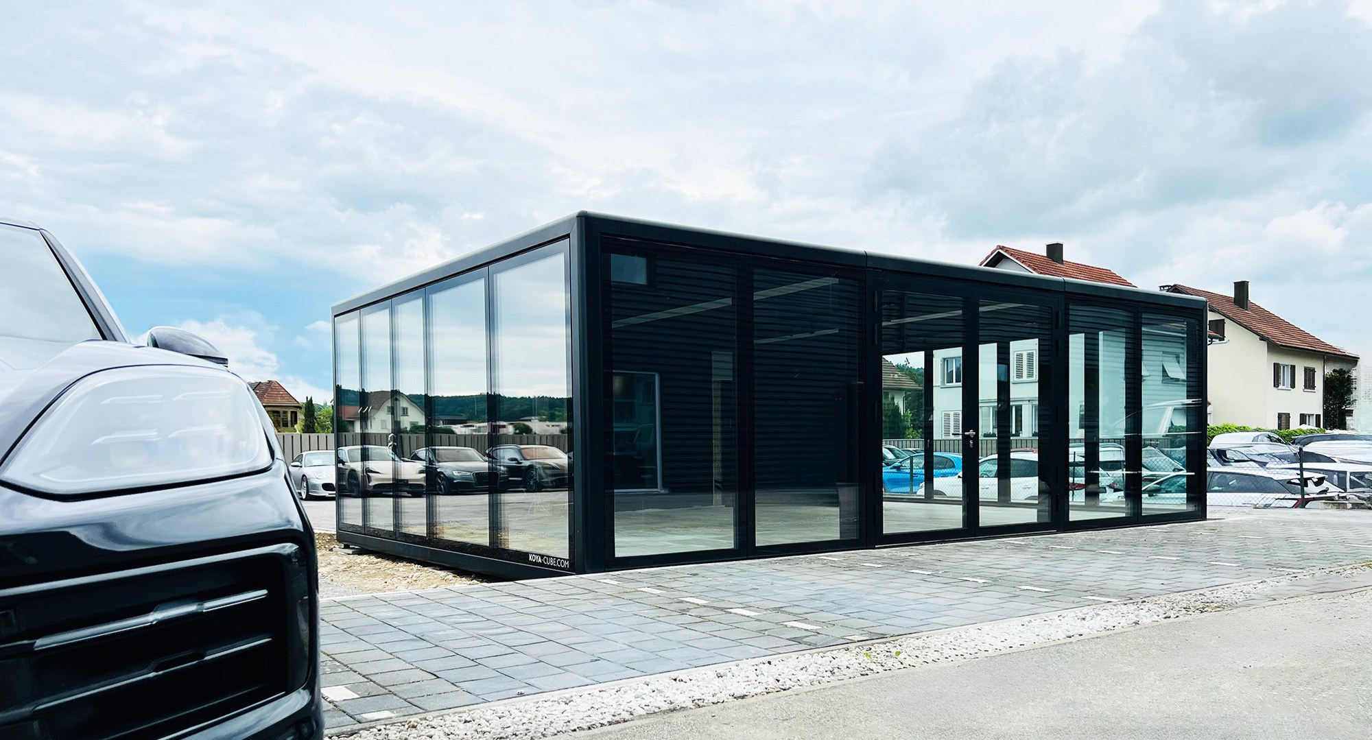 Schwarzer Glaskubus mit Glasfronten, links im Vordergrund die Front eines dunklen Autos, am Sockel KOYA-CUBE.COM.