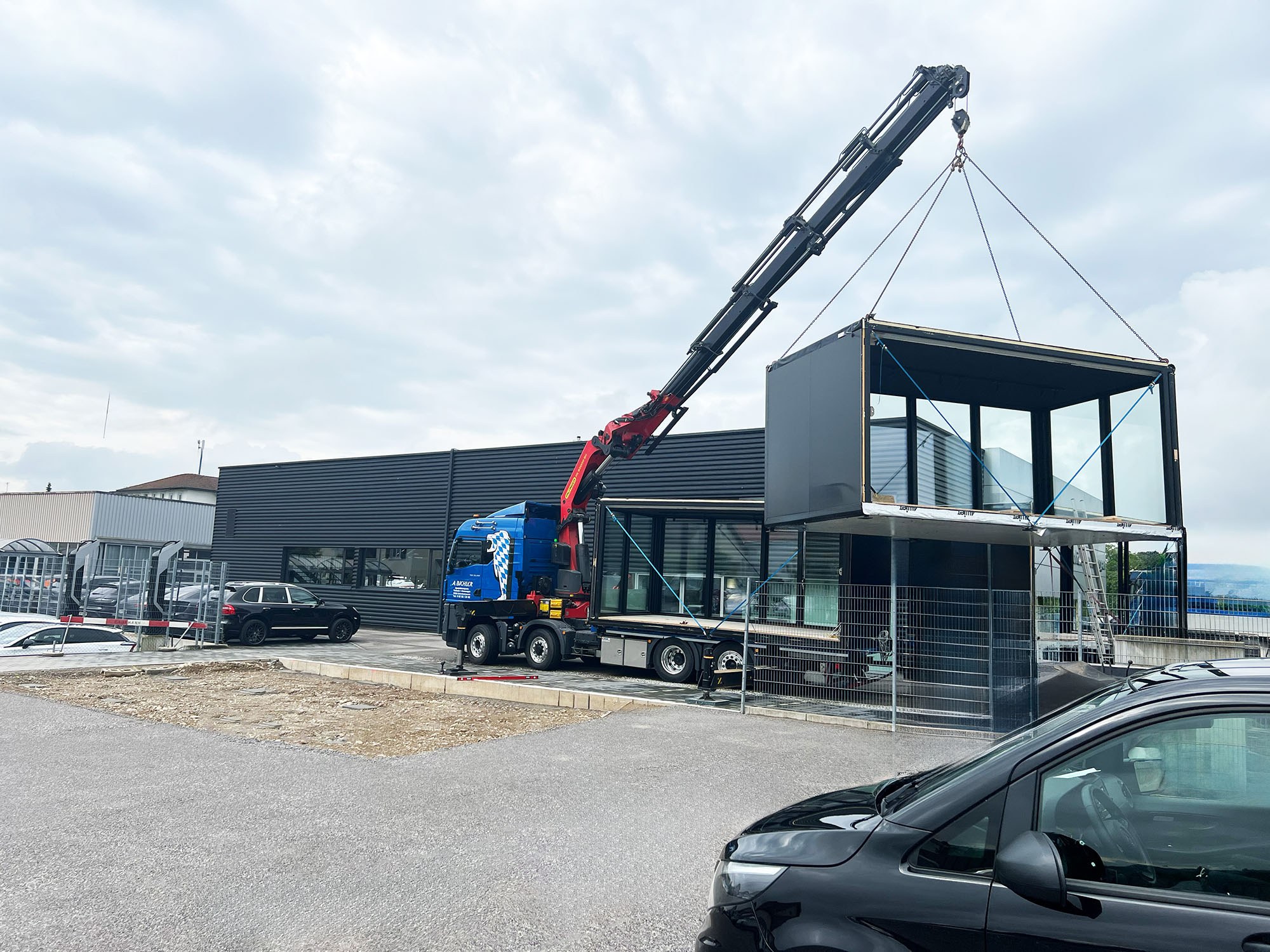 Kran hebt einen schwarzen Glaskubus an Seilen über einen zweiten Glaskubus auf einem Lkw, vorn ein dunkler Pkw.