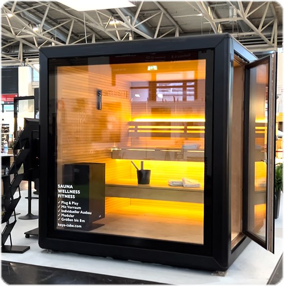 Schwarz gerahmter Glaskubus mit beleuchtetem Saunainnenraum auf einer Messe, auf der Glasfront der Text „Sauna, Wellness, Fitness“.