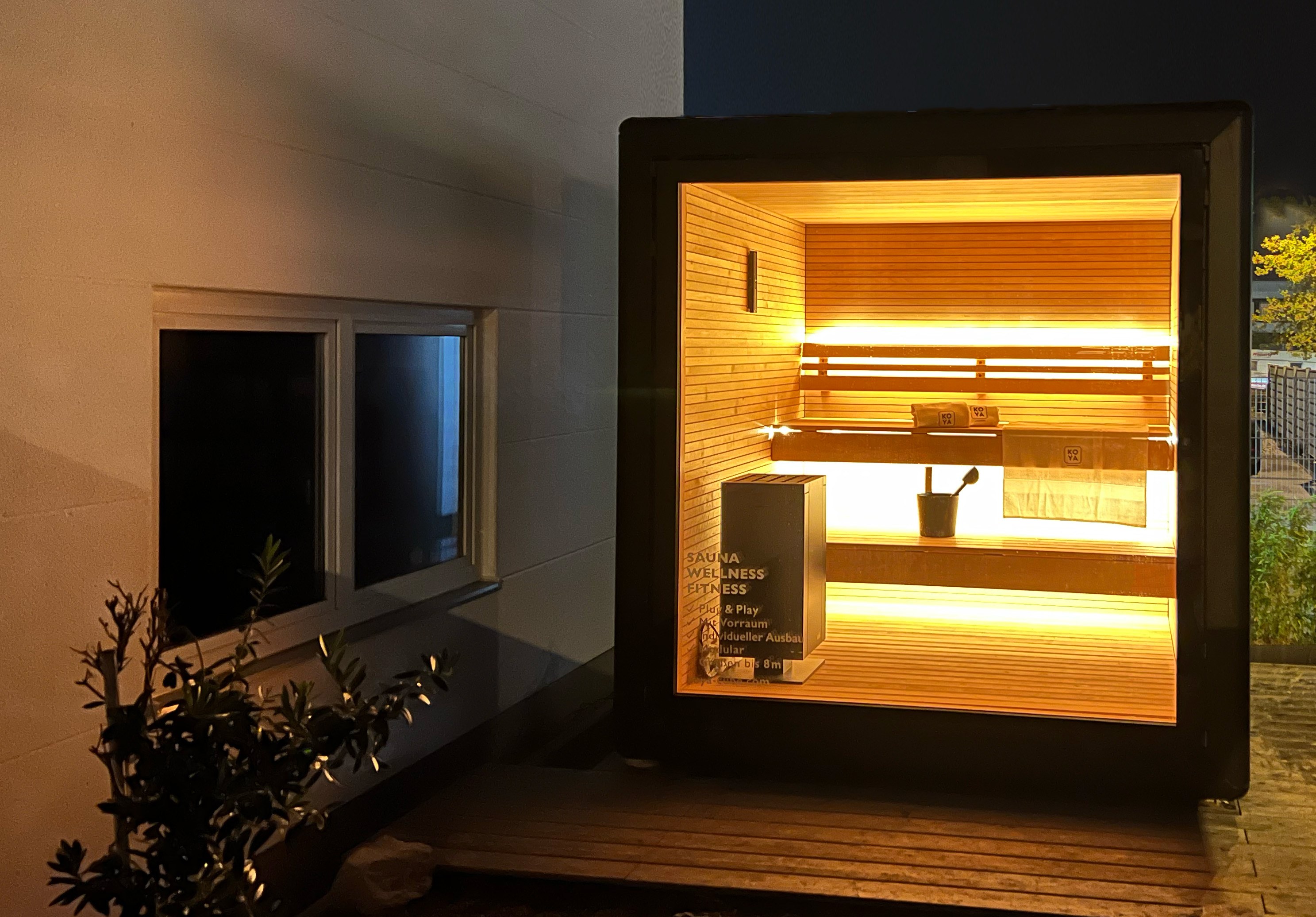 Beleuchtete Saunakabine bei Nacht mit Holzbänken, Ofen, Eimer und Handtuch mit KOYA-Schriftzug, daneben Hauswand mit Fenster.