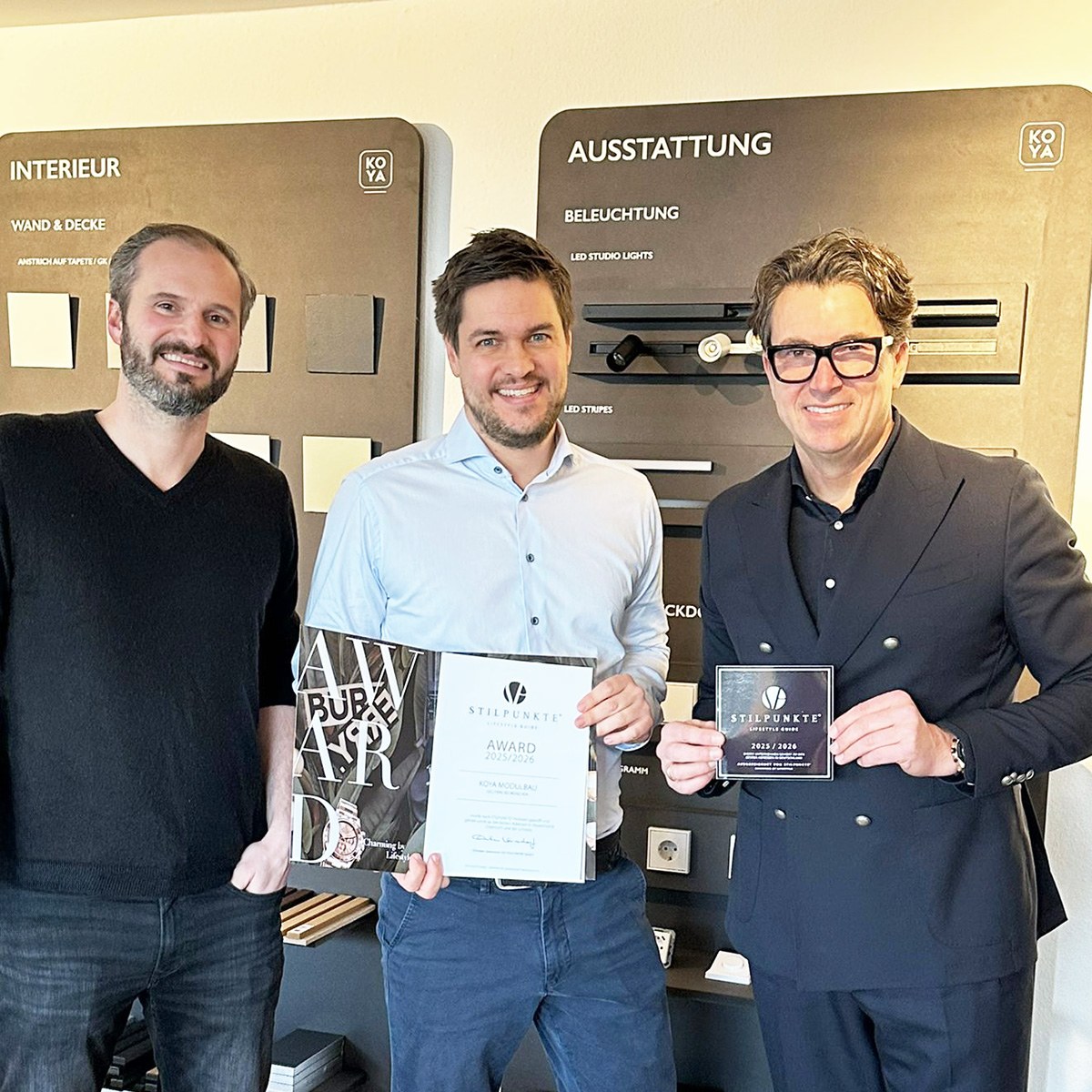 Drei lächelnde Männer vor Showroom-Tafeln; der mittlere hält eine Urkunde „Award 2025/2026“, der rechte eine Plakette.