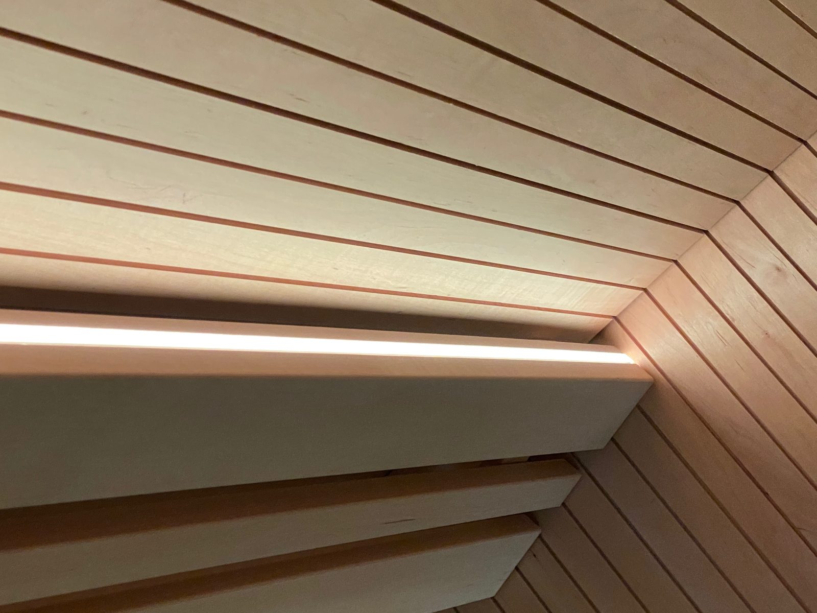 Warmweißes Lichtband an einer Saunabank aus hellem Holz, dahinter waagerechte Wandprofile, aus der Nähe